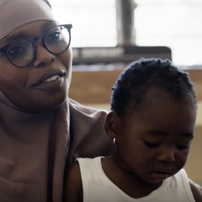 A woman wearing glasses and a headscarf holds a child in her lap. The child is looking down.