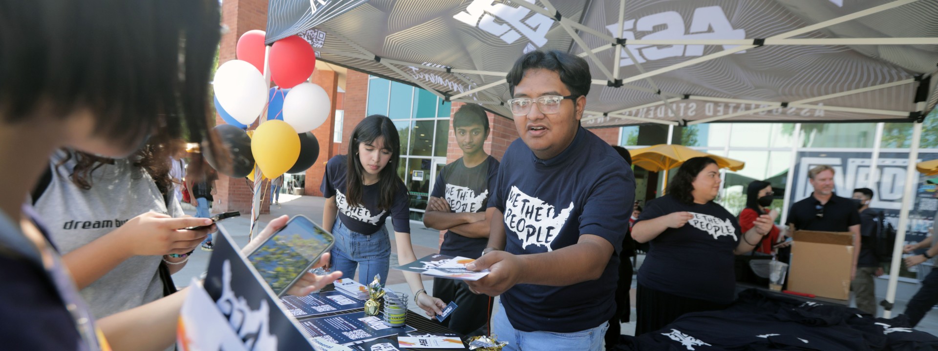 A student wearing a "We the People" t-shirt and standing under an Associated Students Inc., canopy hands students a brochure.