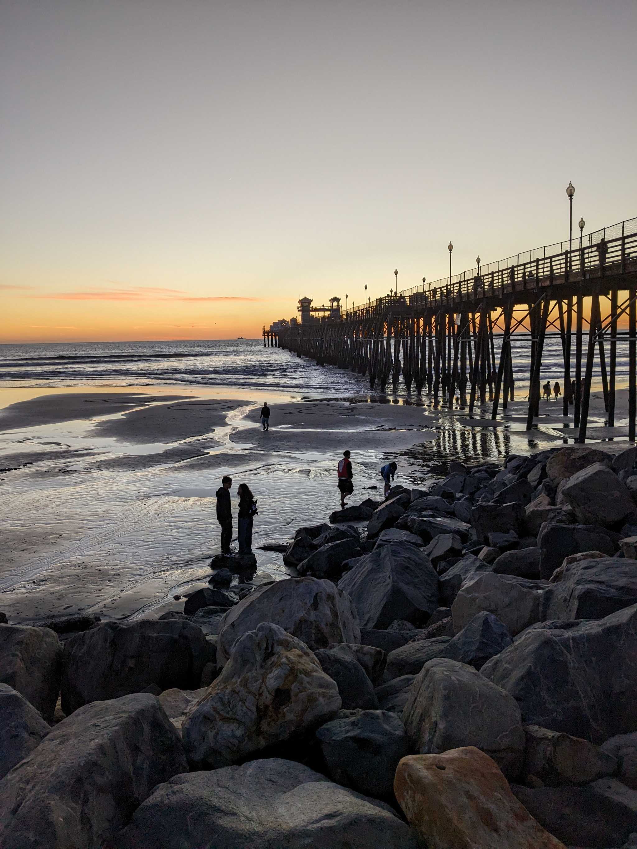 Looking out from below a long ocean pier after sunset with lots of exposed beach and people walking around.