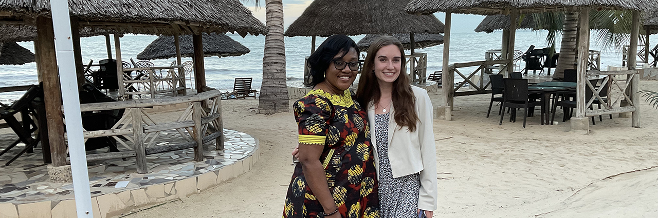 Two women standing by sand, trees, and huts.