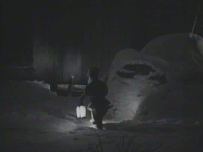 Liten tomte med lykta fotad bakifrån i mörkt snölandskap.