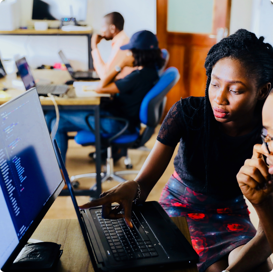 A Black businesswoman assisting a Black colleague by pointing at a laptop screen in an office.