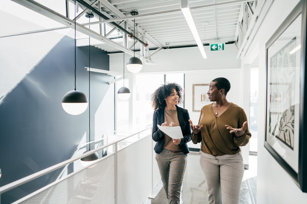 Two female colleagues, one holding a document, walking down an office hallway and chatting.