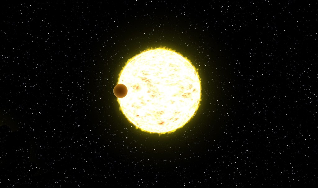 A small brown planet is seen in silhouette crossing in front of a glowing pale yellow star. The background is black with tiny white stars speckled all over.