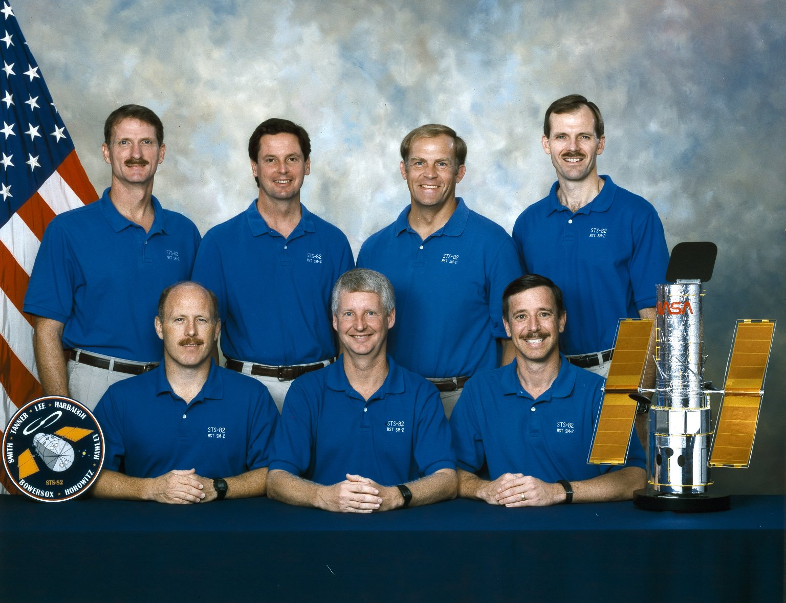 Crew photo with seven astronauts, 4 standing and 3 seated at a table in front of the American flag and a Hubble model on the table.