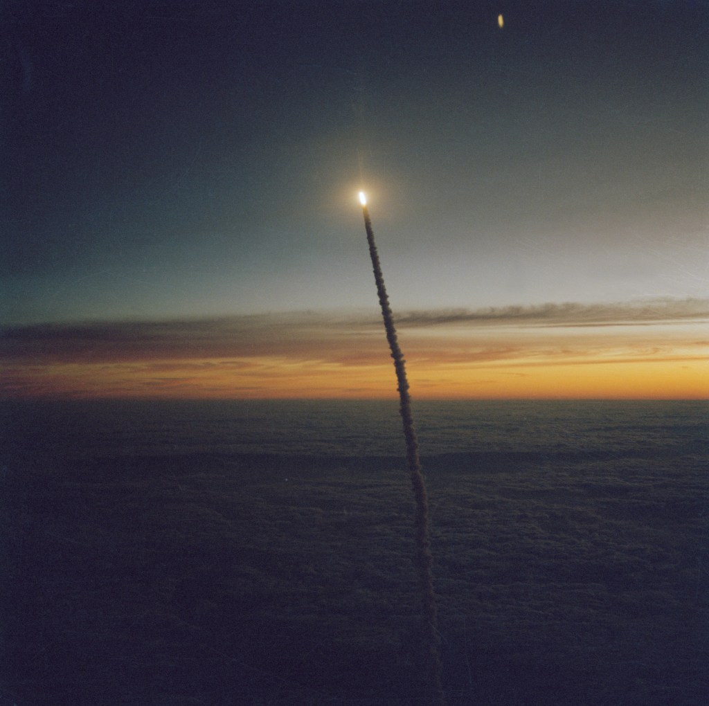 View of the early morning launch of STS 41-G Challenger. The orbiter appears as a bright light at the top of a column of smoke against the sky.