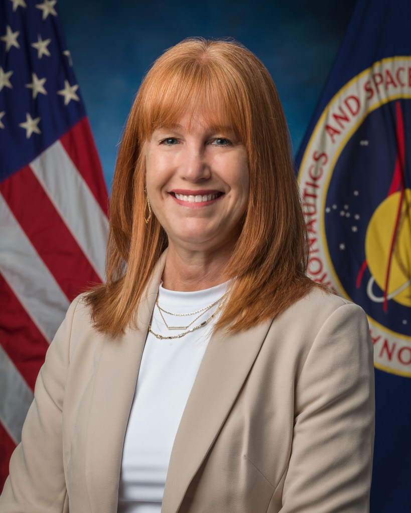 A woman with red hair wearing a white shirt and Khaki blazer poses in front of the United States and NASA flags.