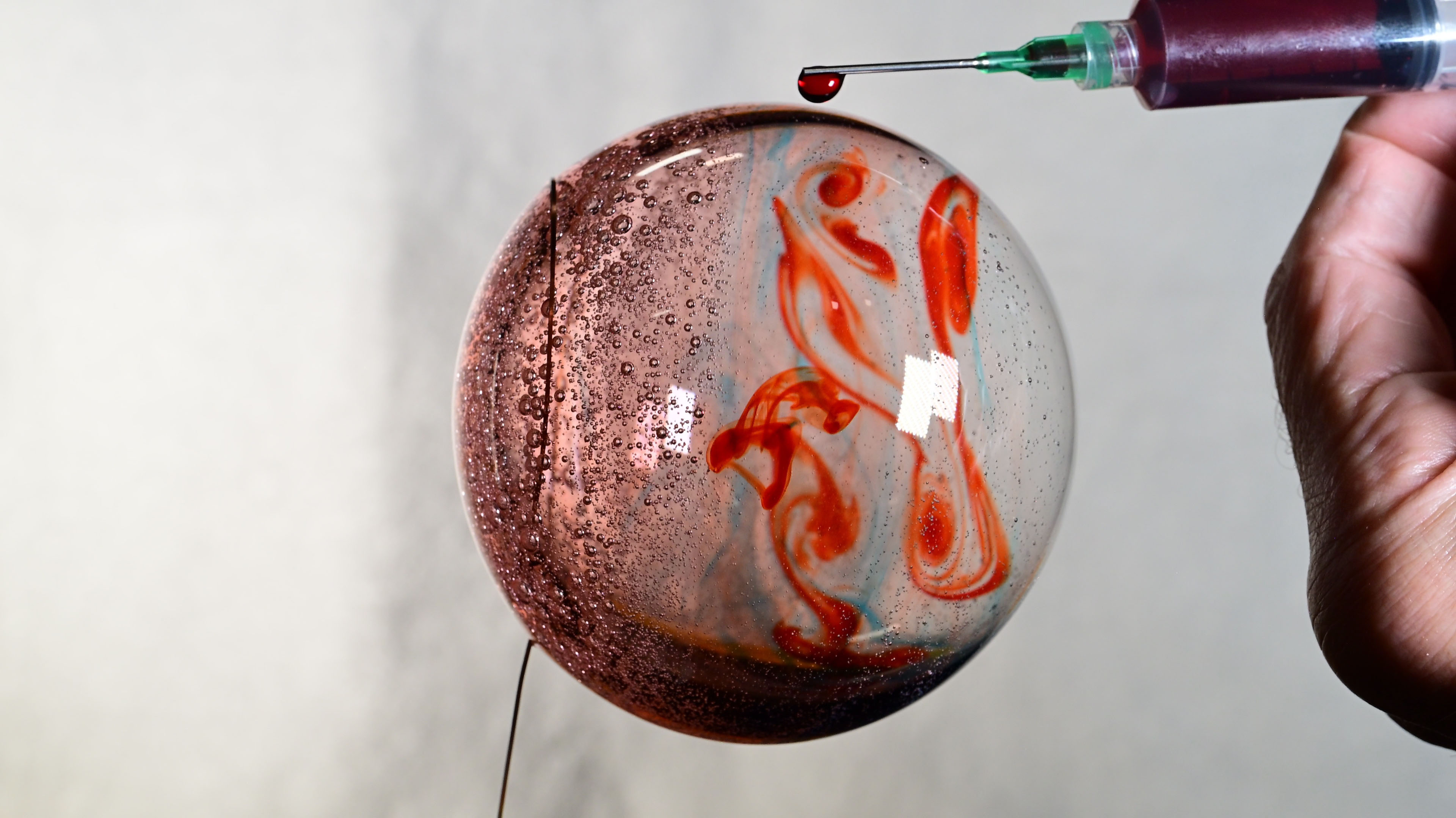 Don Pettit's hand is at the right edge of the photo. He holds a syringe filled with red food coloring. A droplet of food coloring hangs off the needle above a bubble of water. The water sphere is pinkish on its left third. Dye swirls through the remaining two thirds, making it look a little like Jupiter.