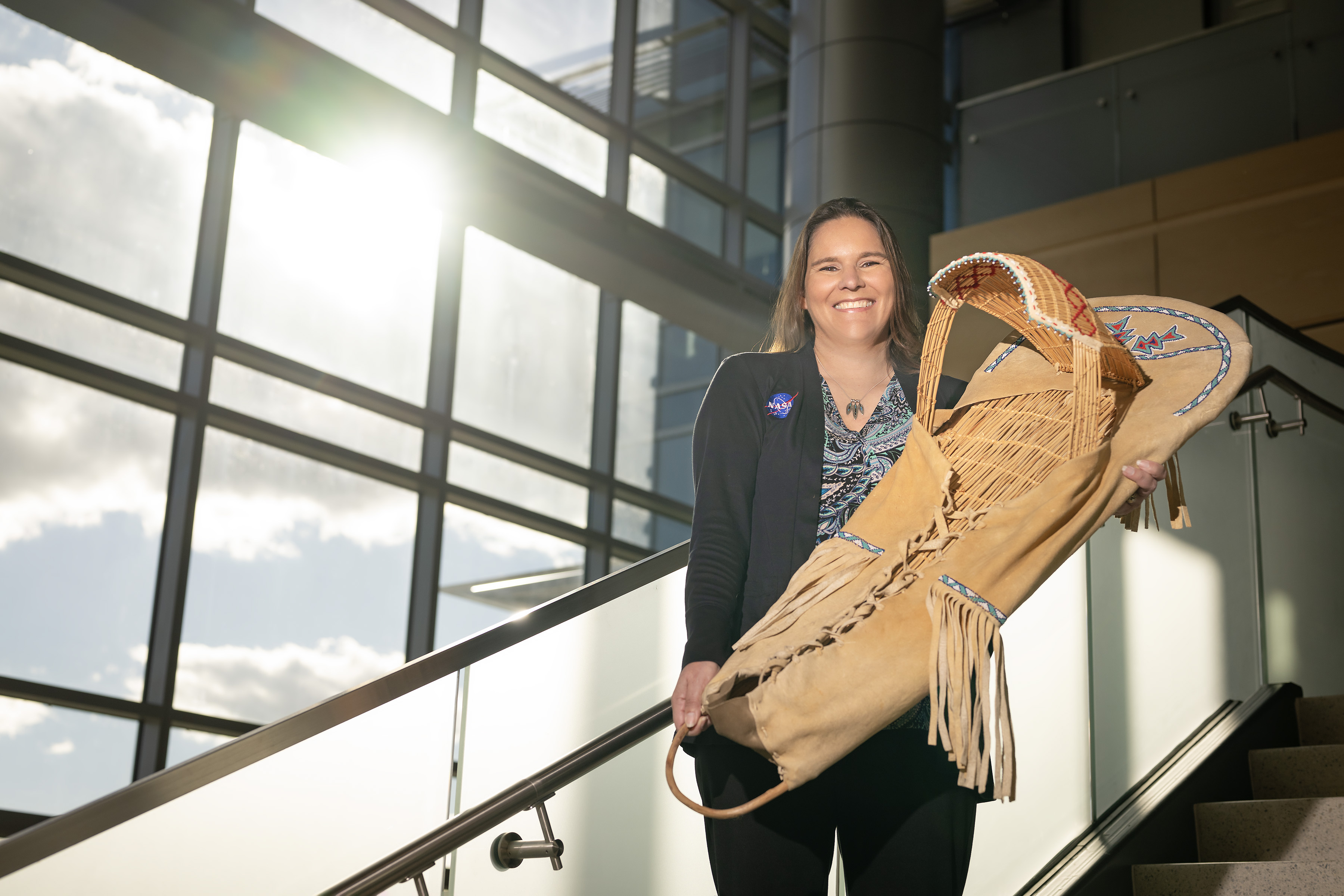 Tami, a woman of Native American heritage from the Delaware Nation, smiles standing on a staircase in a modern, sunlit building, holding a traditional handcrafted cradleboard adorned with intricate beadwork. She is wearing a paisley patterned shirt covered with a black sweater that has the NASA insignia on the right breast.