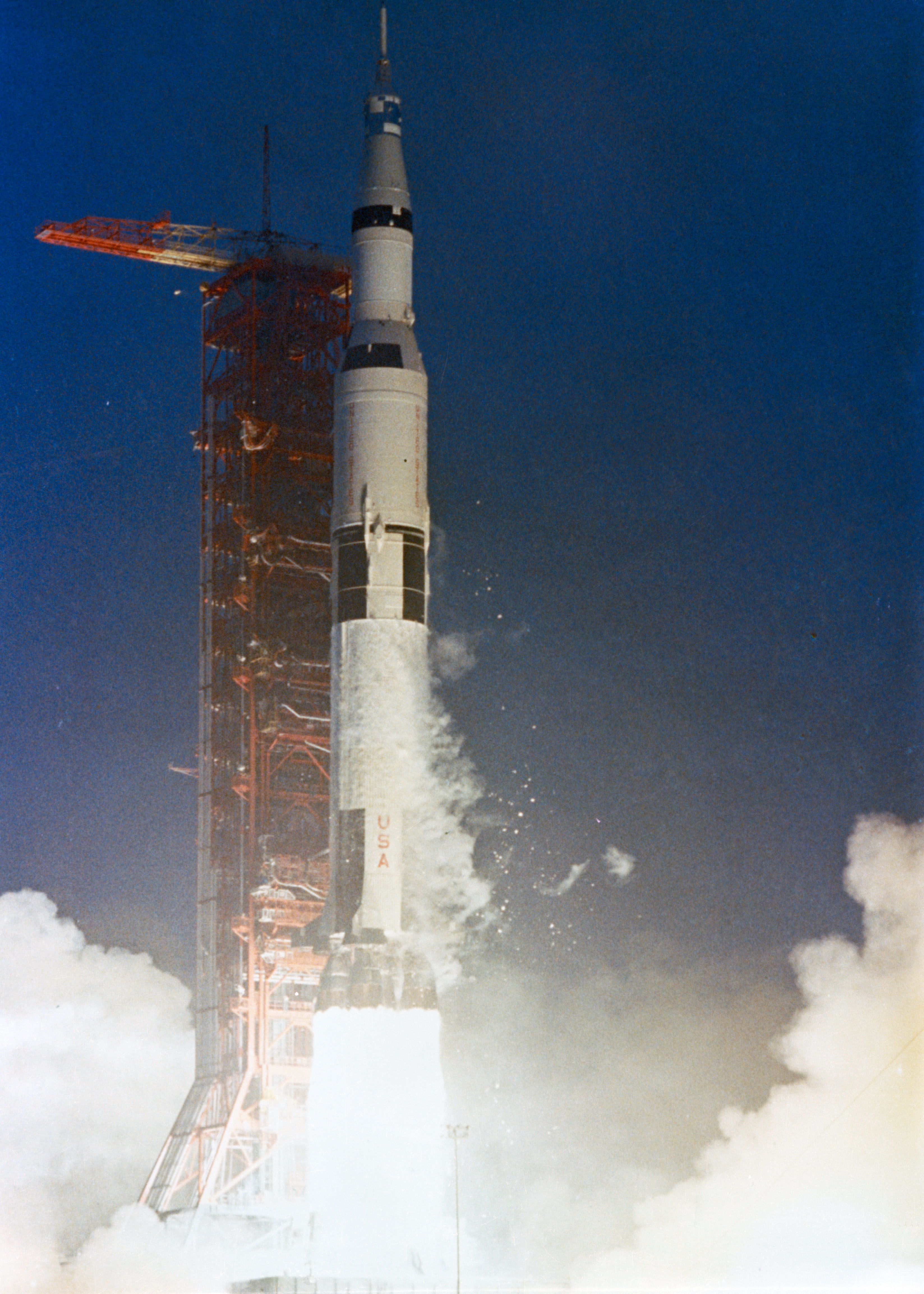 A white rocket lifts off from a launchpad, contrasting against the dark blue sky. A column of flames casts a bright light on the bottom of the image, highlighting white vapor spreading outward. An orange gantry is visible behind the rocket.