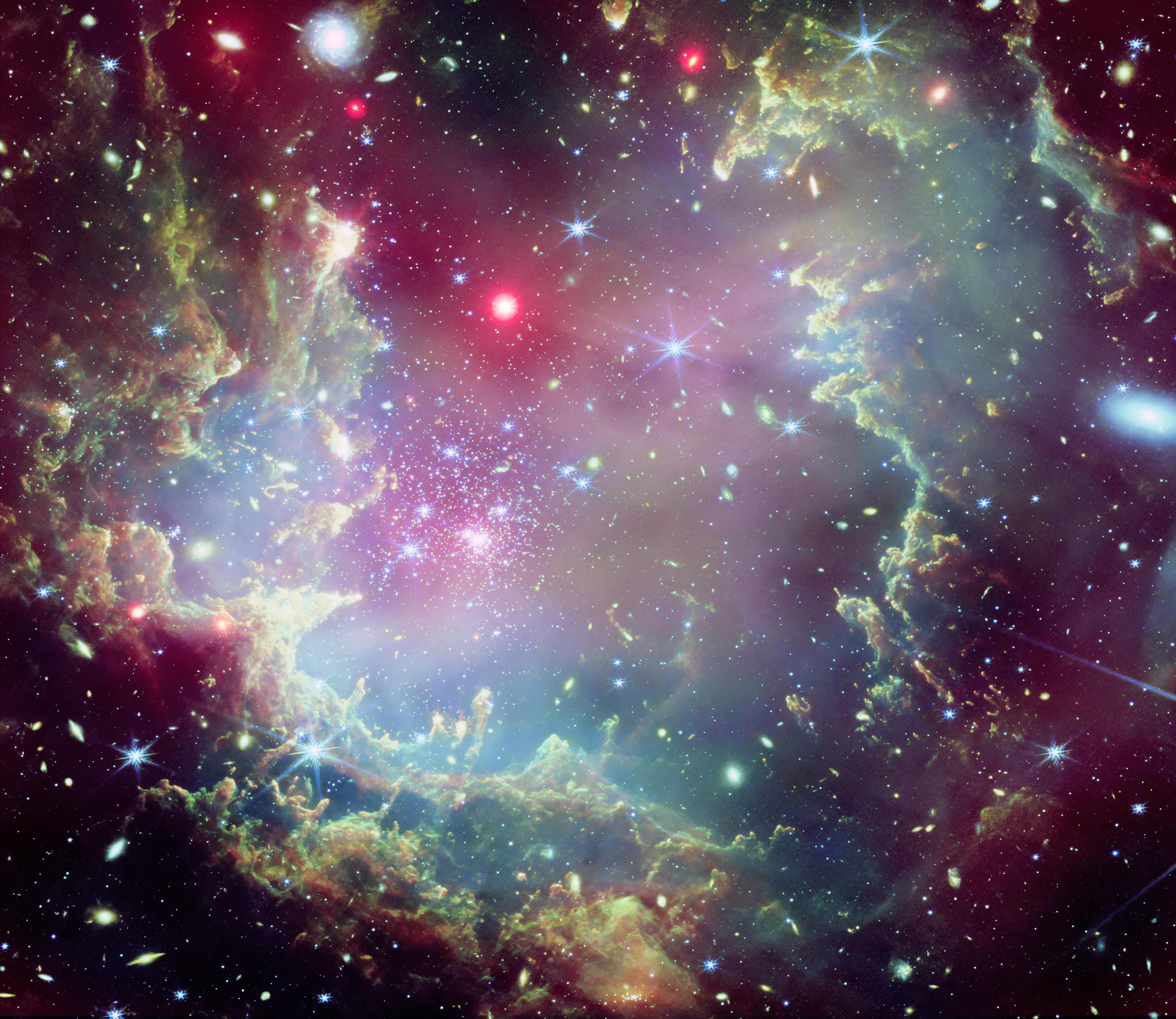 A star cluster is shown inside a large multicolored nebula that looks like a Christmas wreath. The material forms dark ridges and peaks of gas and dust surrounding the cluster. The inner edge is lit. Numerous galaxies are scattered around and within the "wreath." A few large and bright stars are visible as well.