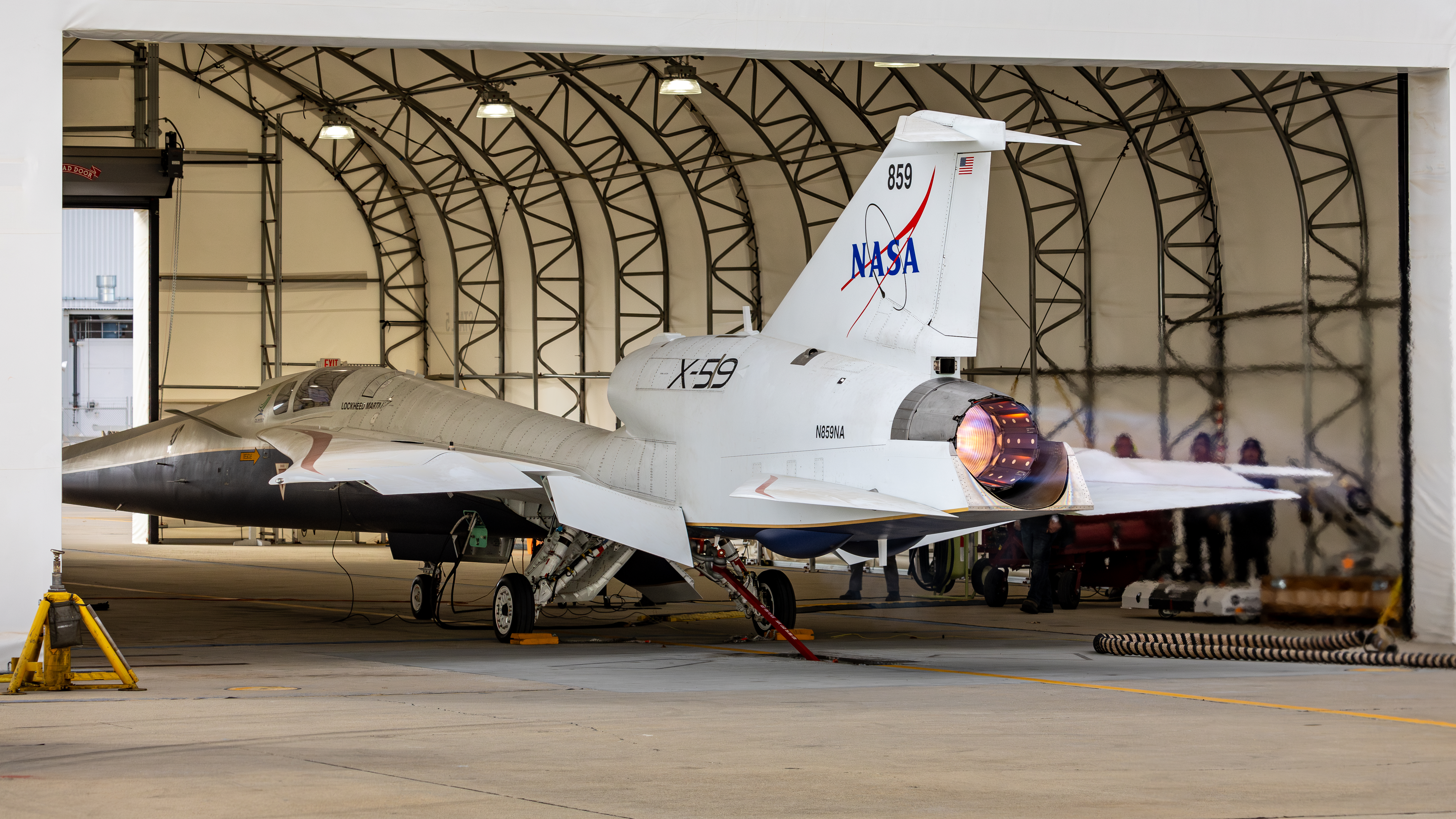 The nearly 100-foot-long X-59 sits partially inside a large run stall, with the rear of the aircraft and its engine extending outside the run stall’s open bay door. Flames are visible from the engine, producing additional thrust during full afterburner tests.