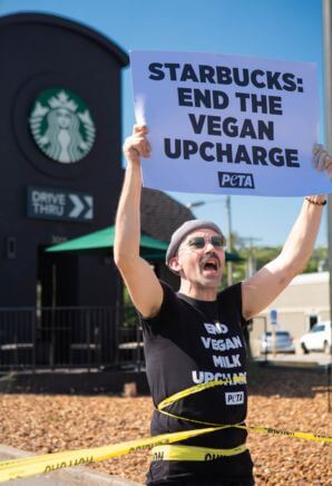 Demonstrator wrapped in police tape holding up a sign in front of a Starbucks that reads "Starbucks: End the Vegan Upcharge!"