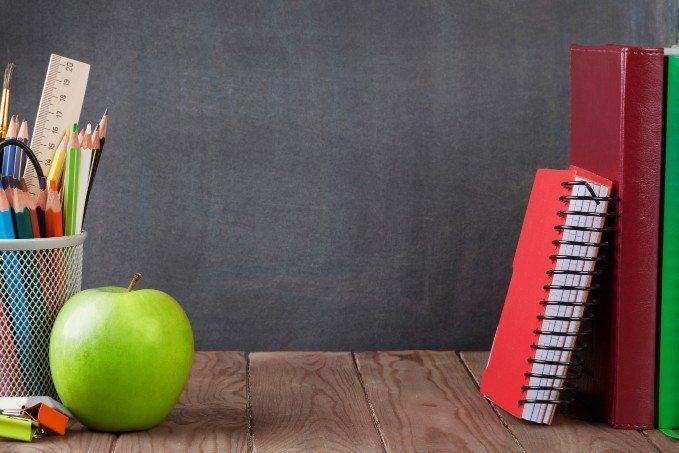 A green apple, colored pencils in a mesh container, a ruler, highlighters, and a red spiral notebook leaning against three upright books on a wooden table with a chalkboard background.