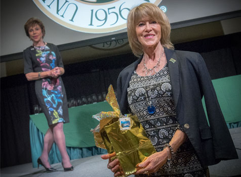 Woman holds a trophy and smiles at the camera, with Dr. Judy Genshaft in the background