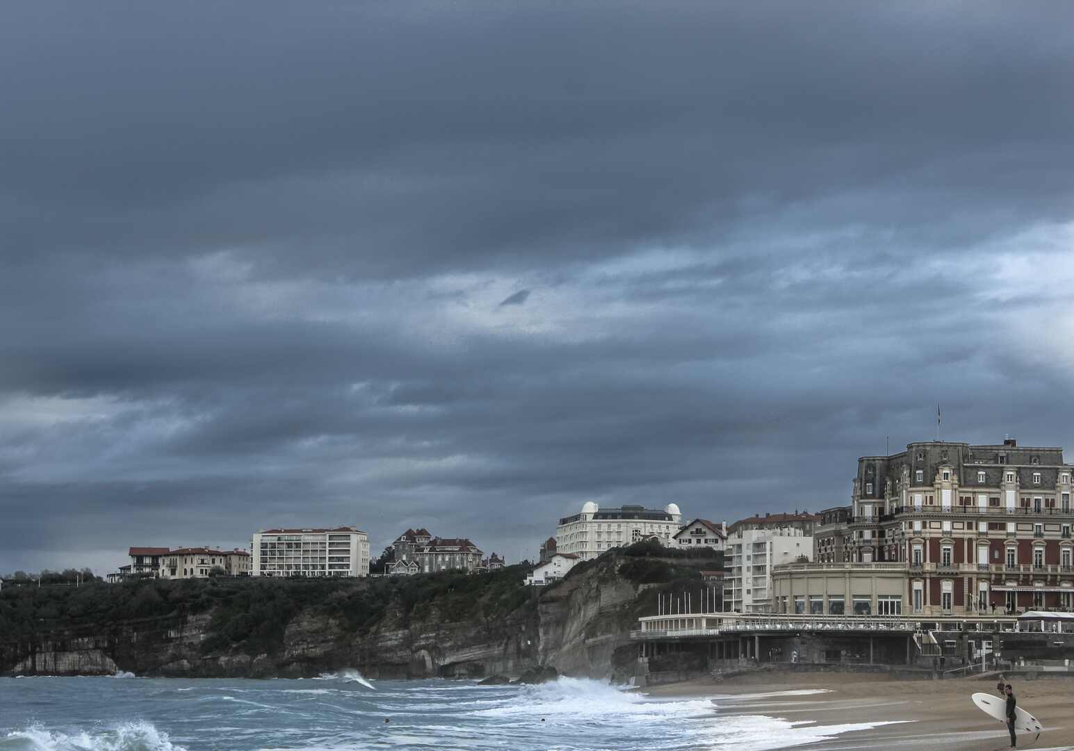 [photo with a beach, cliffs, buildings, dark clouds and a surfer]