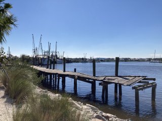 A rickety pier on the water on a clear day with shrimp boats moored in the background.