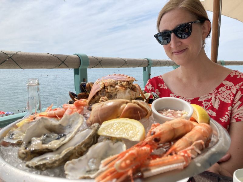 Jessica sitting outdoors with the sea in the background and a big seafood platter in the foreground, filled with crab, prawns and oysters.