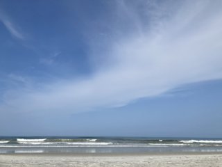 A sandy beach with gentle waves crashing under a blue sky with wisps of cloud.