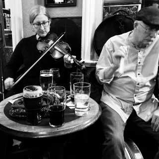 A woman playing fiddle and a man playing bones at a pub table covered with pints.