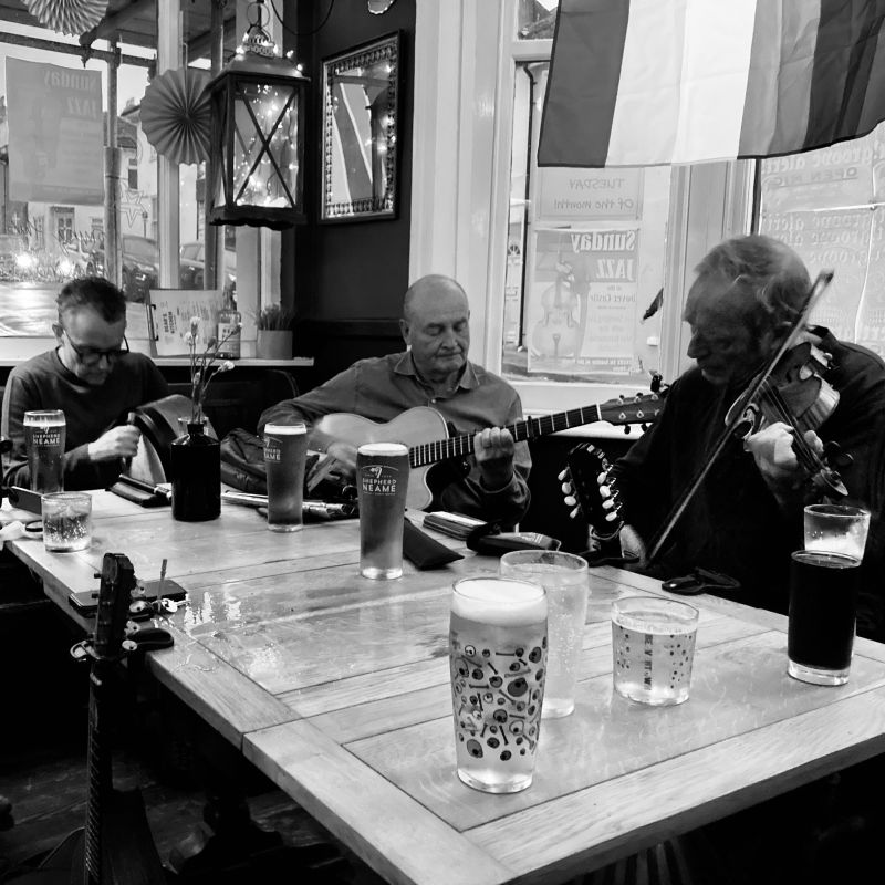 A bodhrán player, guitarist and fiddler playing around a pub table with drinks on it.