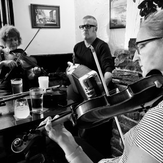 Two fiddlers and an accordion player gathered round a pub table, playing.