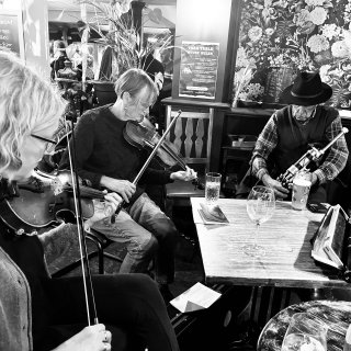 Two women playing fiddle and a man in a hat playing pipes in a pub with ornate wallpaper.