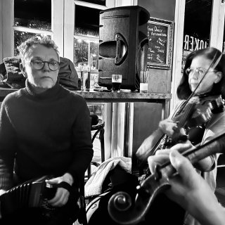 A man playing concertina and a woman playing fiddle with a hand holding another fiddle in the foreground.