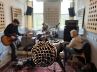 Close up of a microphone. In the background, musicians are sorting out their instruments: guitar, drums, and bass.