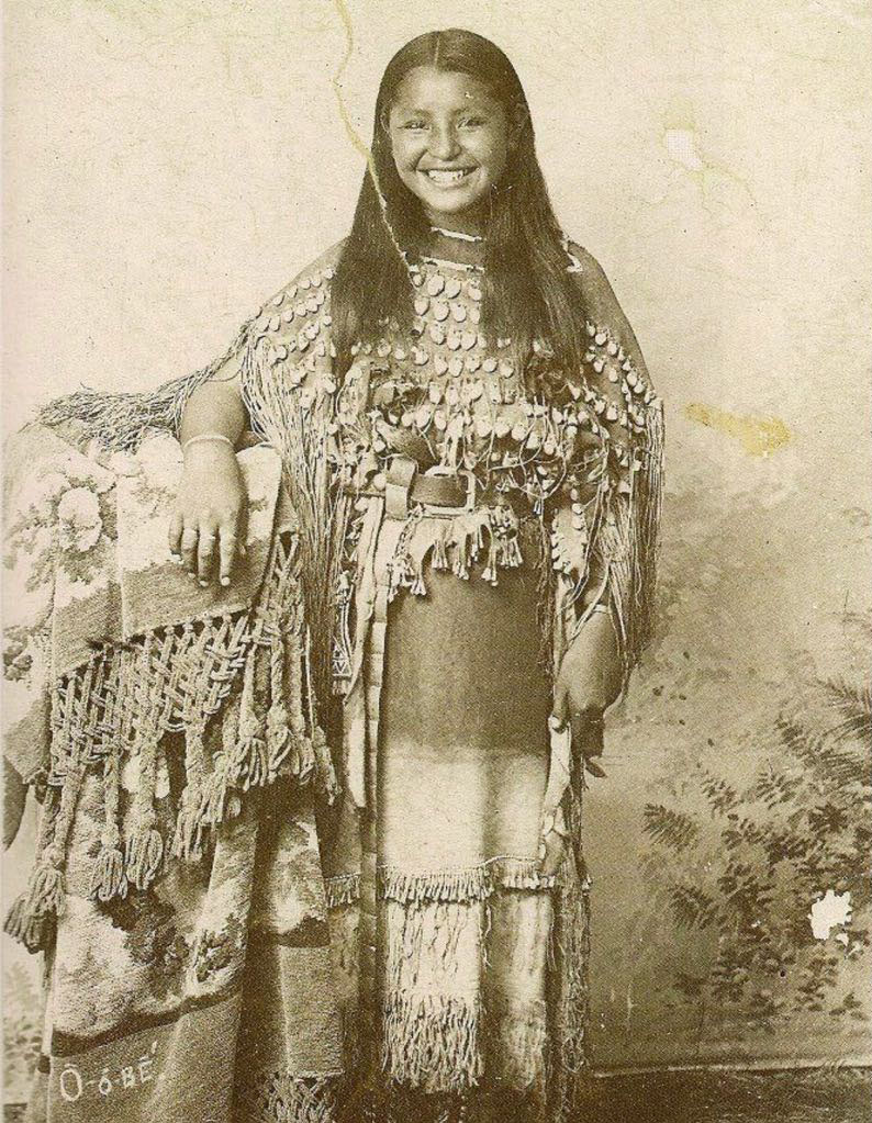 O-o-dee of the Kiowa tribe in traditional dress with a heartwarming smile on her face in a photograph over 100 years old.