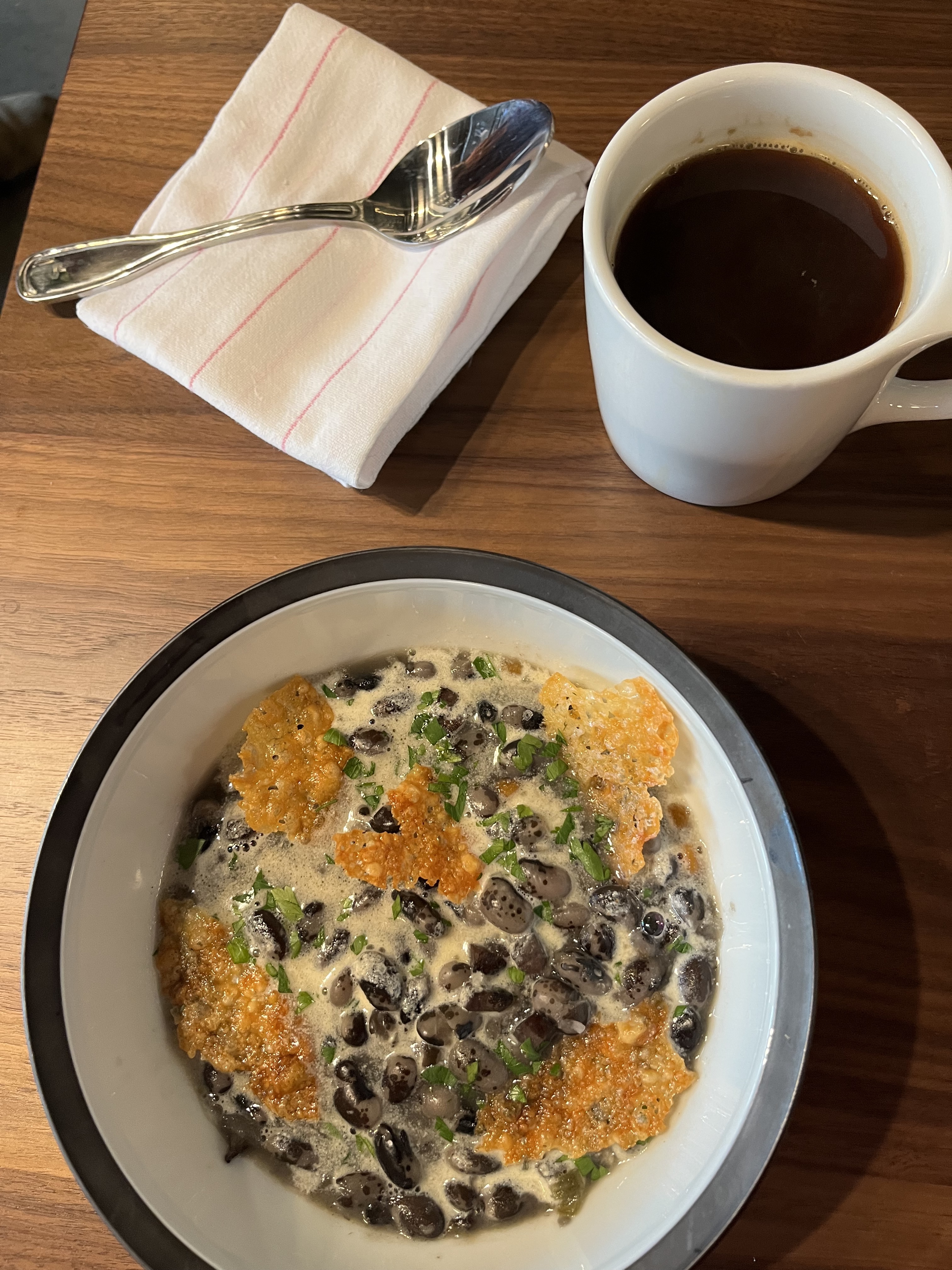 A bowl of bean soup on a table next to a coffee.