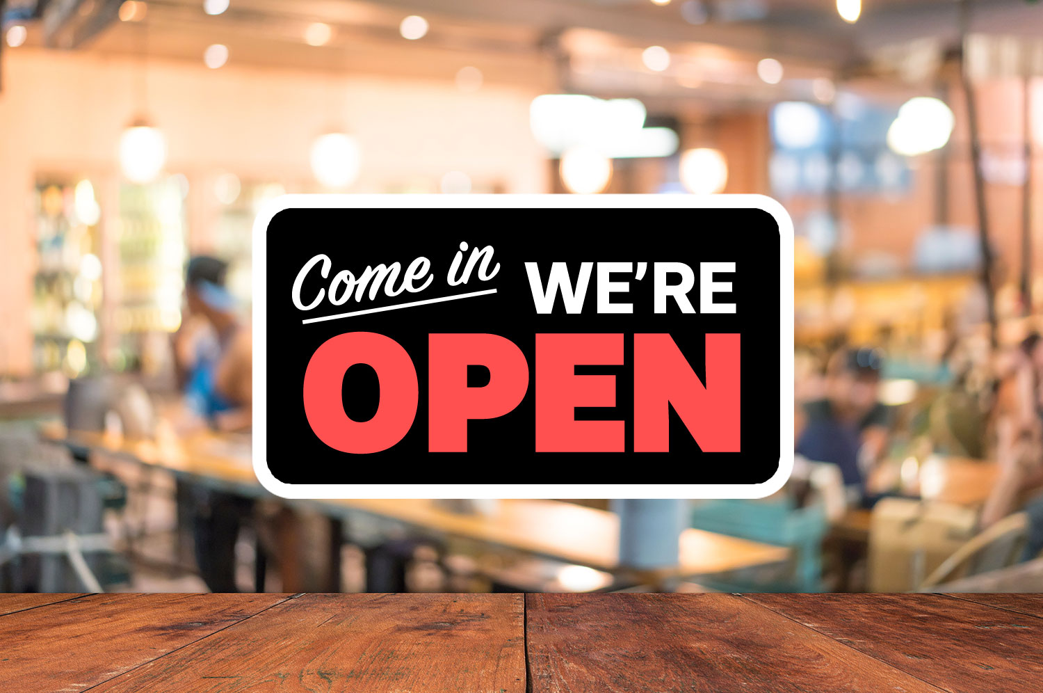 A stock photo of a now open sign with an out of focus dining room visible behind it 