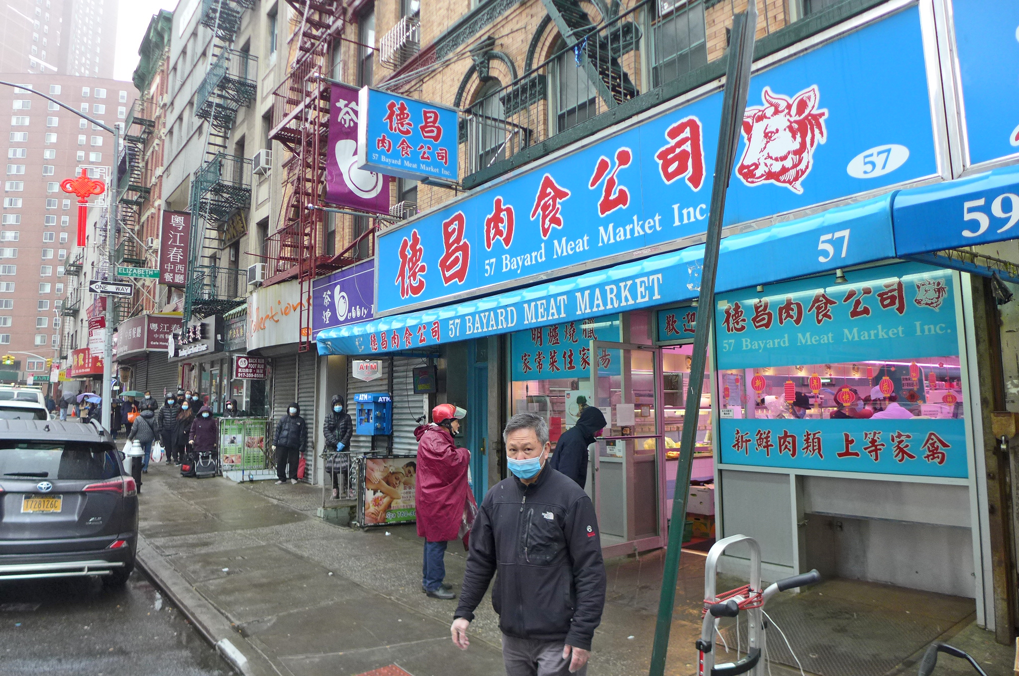 A supermarket with a blue awning, a man in a surgical mask stands in the foreground.