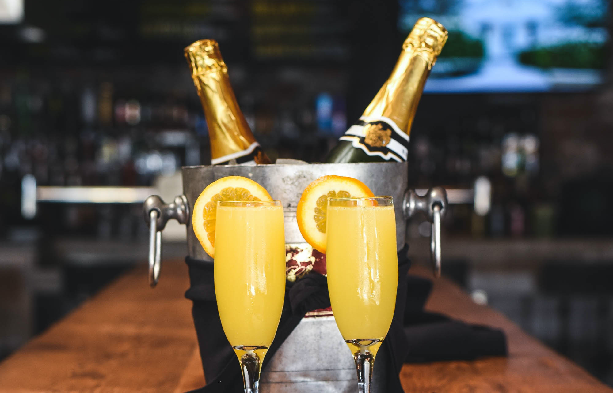 Two champagne bottles in a icy chiller flanked by mimosas with orange wedges.