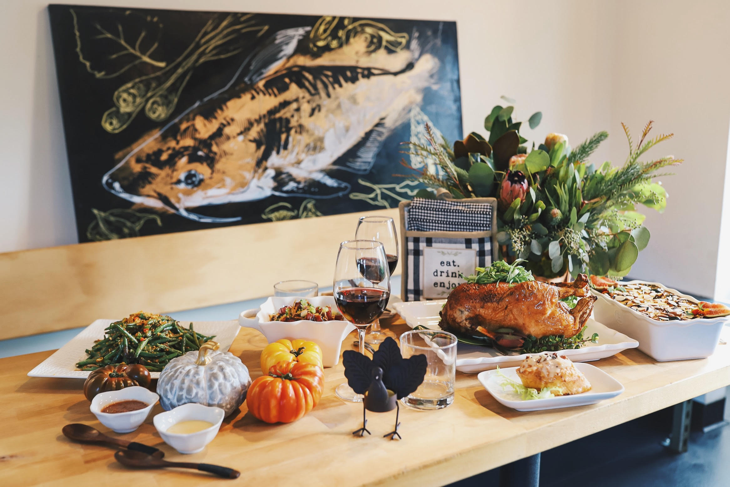 A table covered with a roasted duck, gourds, sauces in tureens, a turkey figurine, and two glasses of red wine, with a painting of a catfish in the background