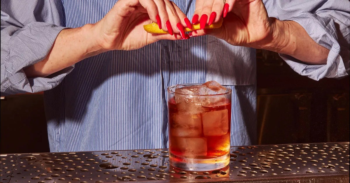 A person with red nails squeezes a lemon twist over a cocktail