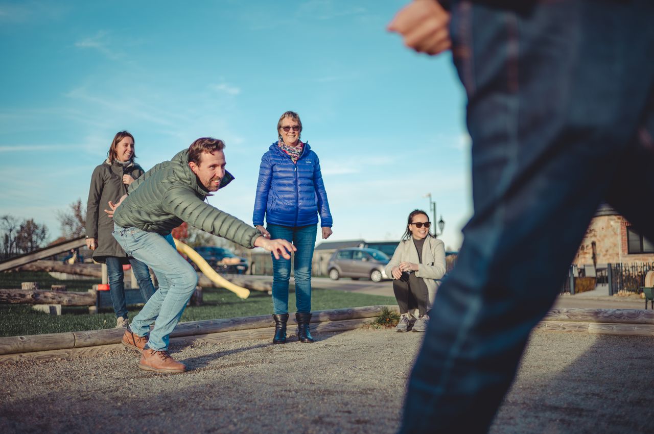 A man tosses a ball with precision as he plays a game of jeu de boules, surrounded by a group of onlookers who smile and laugh at the friendly competition.