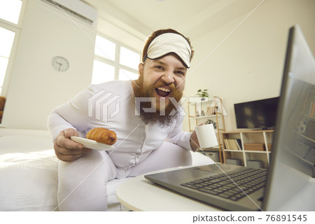 Happy fat man in pajamas sitting on bed in the morning, enjoying breakfast and laughing 76891545