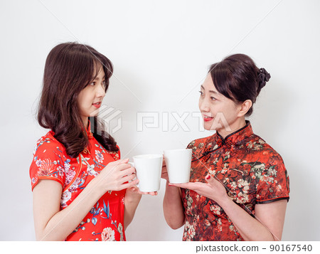 Portrait of mature asian mother and her asian daughter both wearing traditional cheongsam qipao dress relaxing drinking on white background. 90167540