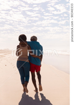 Rear view of african american couple with arms around walking at sandy beach against cloudy sky 90340333