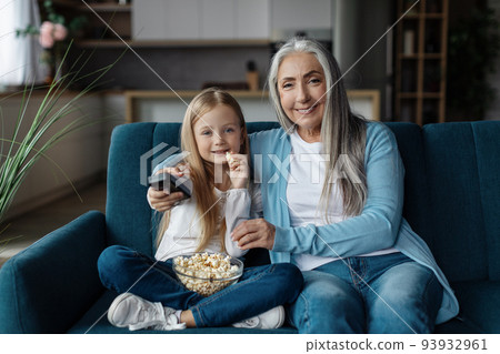 Glad european little girl and mature grandmother with remote control eat popcorn and watch tv 93932961