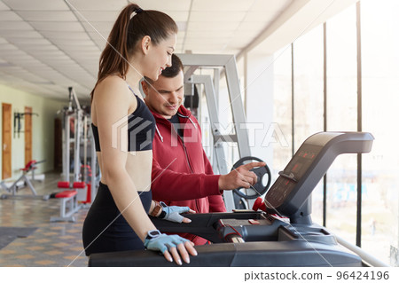 Cheerful magnetic young lady asking for help her trainer, trying to use running machine, listening to instructions carefully. Attractive girl wears black tight top, shorts and light blue gloves. 96424196