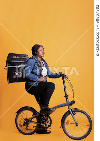 Restaurant courier holding thermal backpack delivering takeaway food order to client during lunch time, standing over yellow background. Pizzeria delivery worker riding bike to deliver fast food meal 98867982