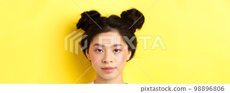 Head portrait of stylish asian girl with bright makeup and hairbuns, looking at camera, standing on yellow background 98896806