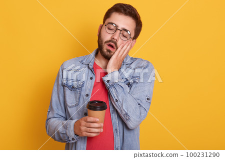 Close up portrait of man over isolated yellow studio background, taking coffee in takeaway paper cup, posing with closed eyes and open mouth, keeps hand on cheek, wearing red shirt and denim jacket. 100231290