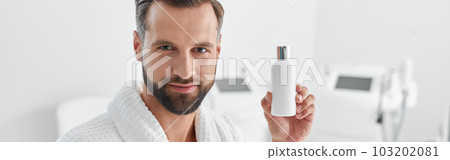 Attractive mature man holds bottle of cosmetic product standing in cosmetologycal clinic 103202081