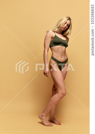 Full-length image of young blonde woman with slim, fit, muscular body posing in green underwear a against yellow studio background 103266683