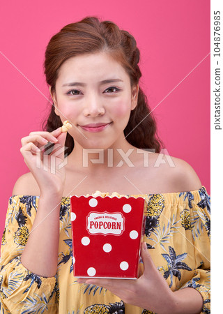 female model wearing a flower-patterned dress with a pink background and eating popcorn deliciously 104876985