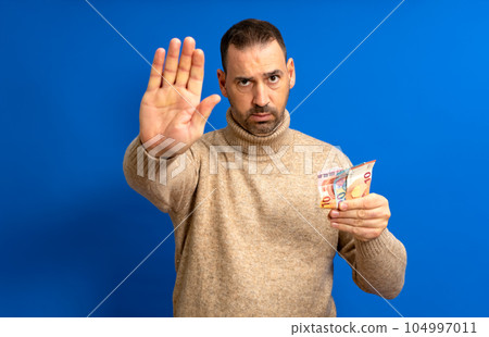 Bearded hispanic man in his 40s wearing a beige turtleneck holding several euro bills and making the stop sign isolated over blue background 104997011