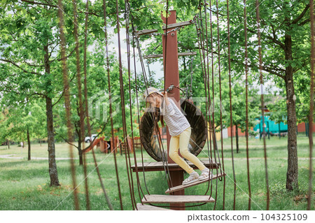 Full length portrait of happy smiling little female child wearing casual clothing and cap playing on rope swings at sunny playground in summer. 104325109